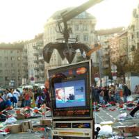 Einarmiger Bandit auf einem müllübersäten und abgesperrten Platz, Menschen im Hintergrund. Eine Kranzange macht den Anschein, den einarmigen Bandit hochheben zu wollen.