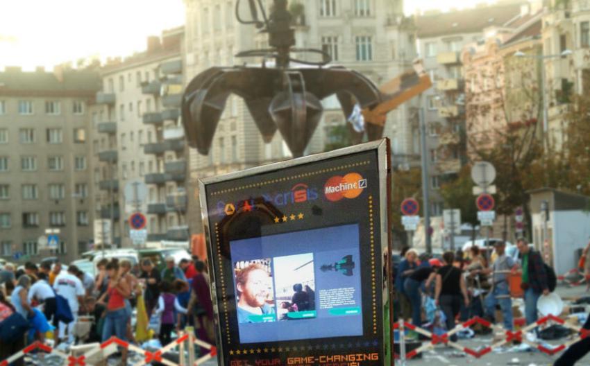 Einarmiger Bandit auf einem müllübersäten und abgesperrten Platz, Menschen im Hintergrund. Eine Kranzange macht den Anschein, den einarmigen Bandit hochheben zu wollen.